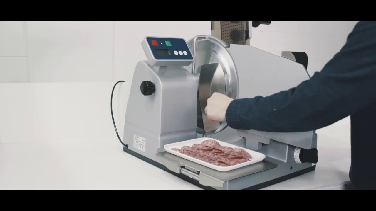 Meat slicer slicing salami. Person operating machine. Slices on tray. Silver machine.