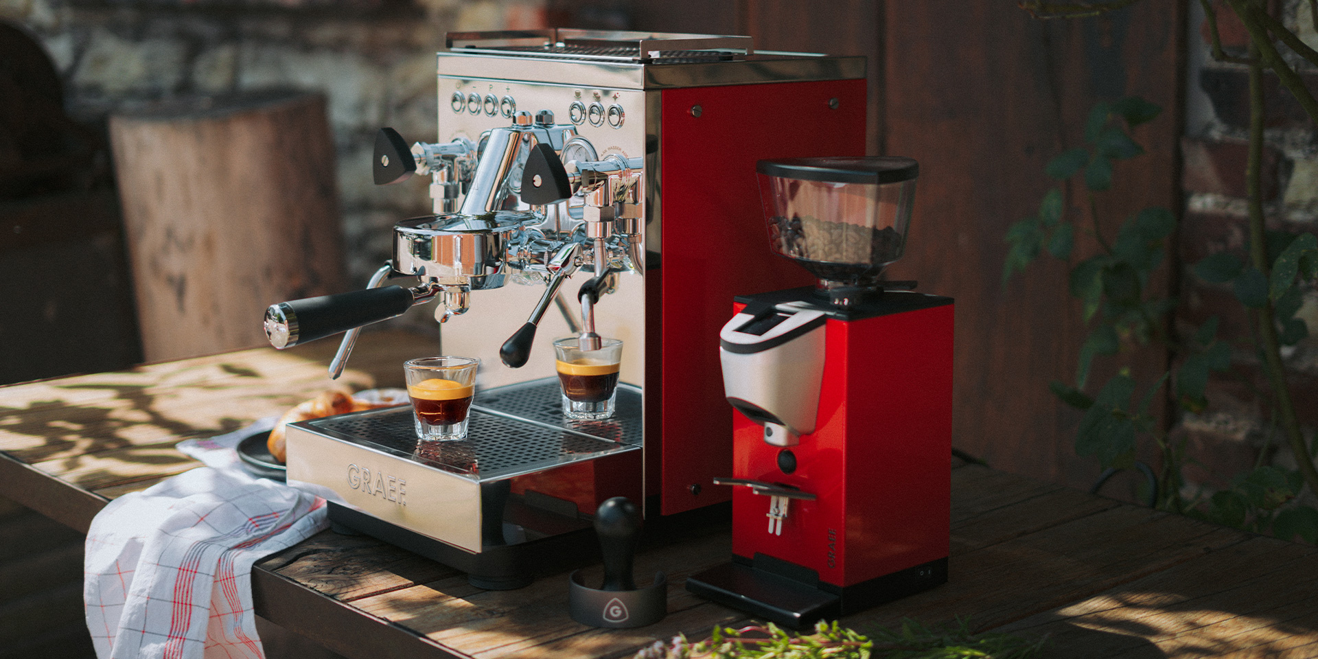 Eine Espressomaschine und ein Kaffeevollautomat stehen auf einem Holztisch im Freien.