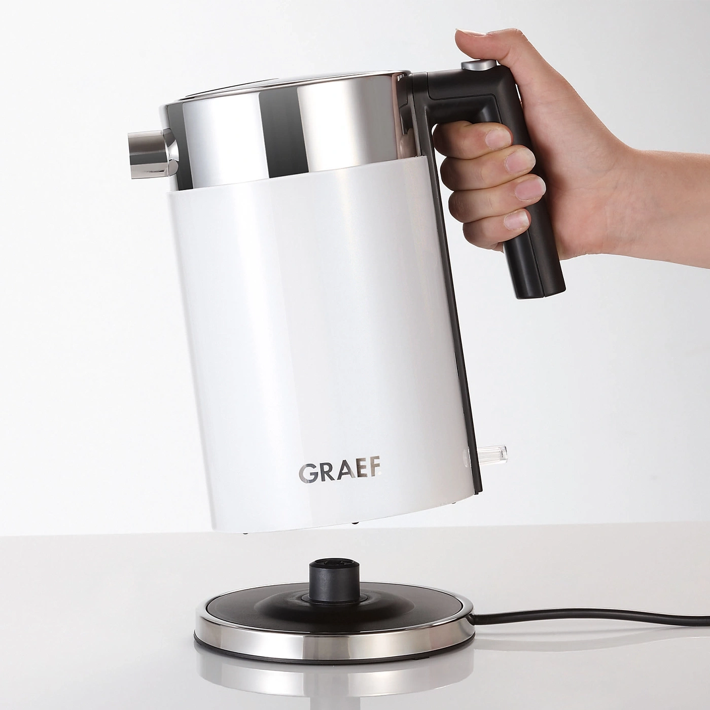 Weißer Wasserkocher mit Griff, der von einer Hand gehalten wird, über einer Basis.