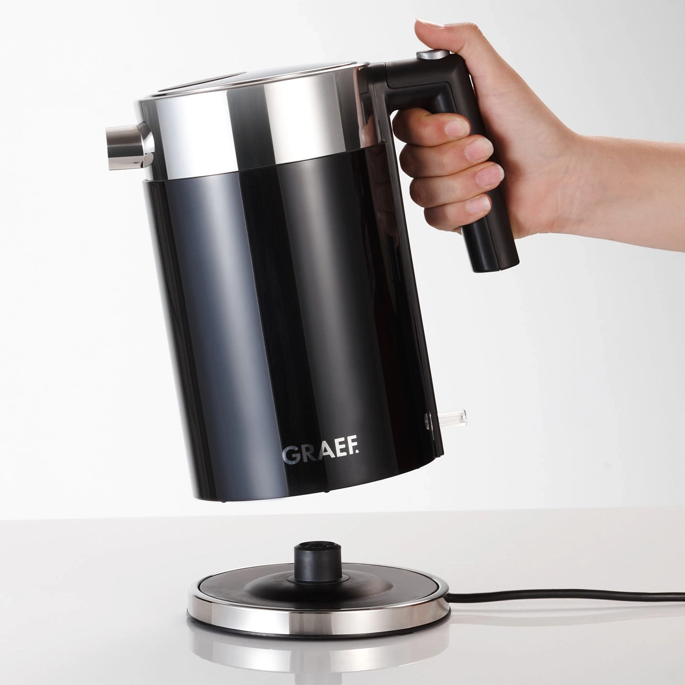 Schwarzer Wasserkocher mit Griff, der von einer Hand gehalten wird, auf weißem Hintergrund.