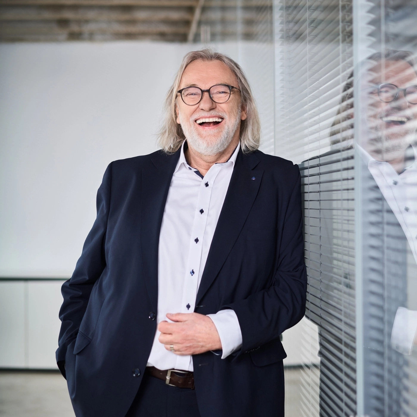Ein lächelnder Mann mit Brille und Anzug lehnt an einer Glaswand.