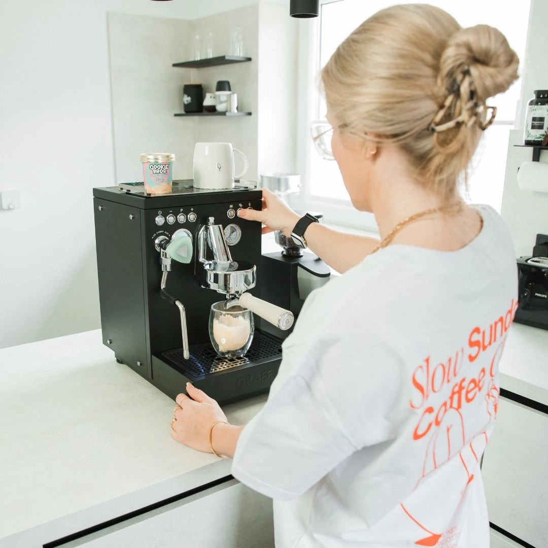 Frau bereitet Kaffee zu. Espresso-Maschine, Tasse, Küche, blondes Haar, Brille, Shirt.