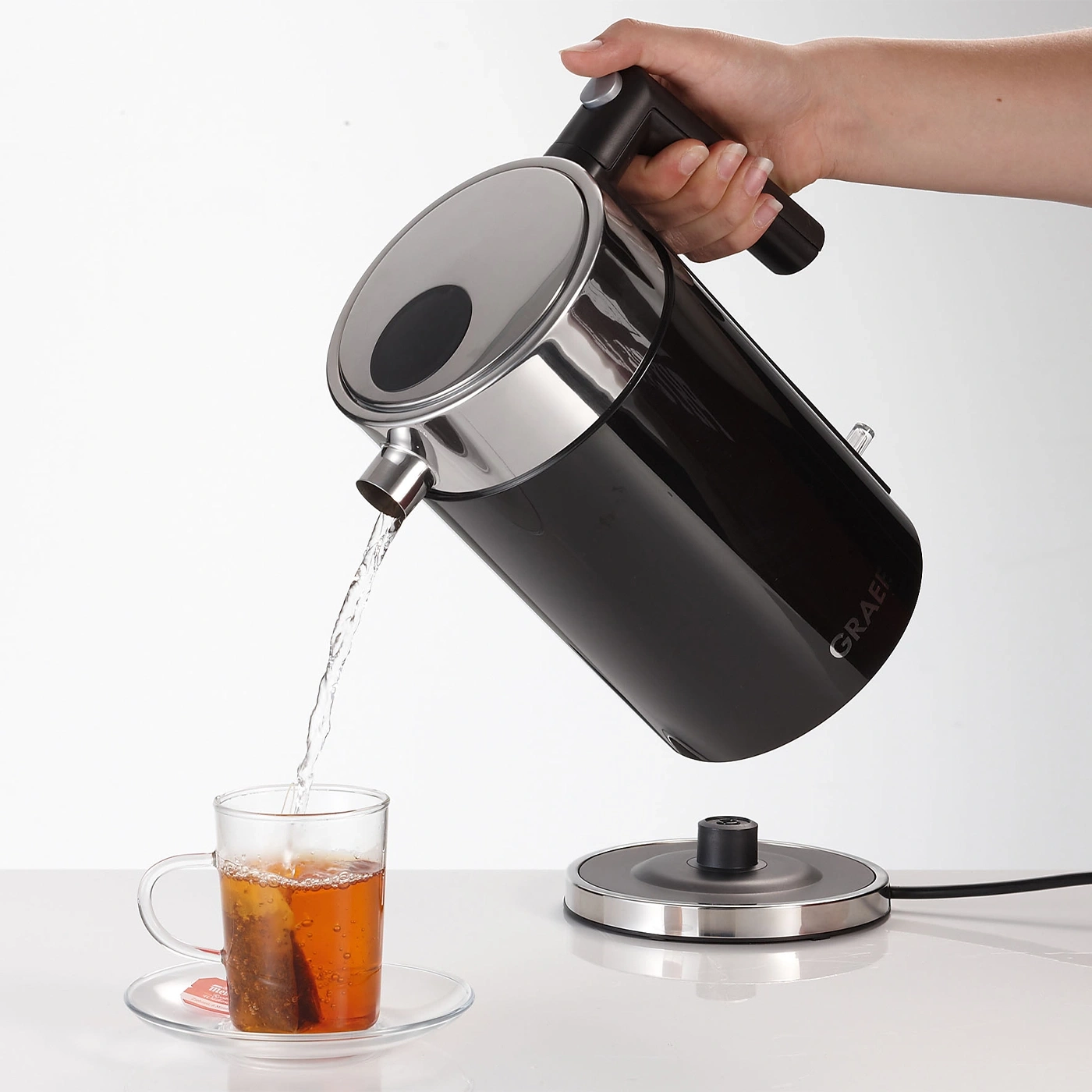 Eine Hand gießt Wasser aus einem schwarzen Wasserkocher in eine Teetasse.