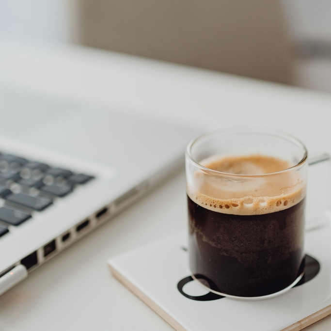 Kaffee in einem Glas auf einem Untersetzer neben einem Laptop auf einem weißen Tisch