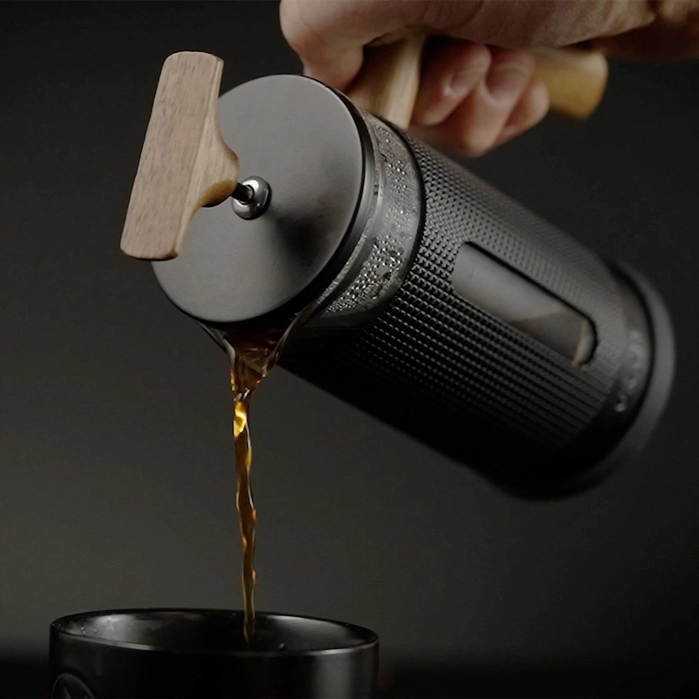 Eine Hand hält eine Kaffeepresse, aus der Kaffee in eine Tasse gegossen wird.