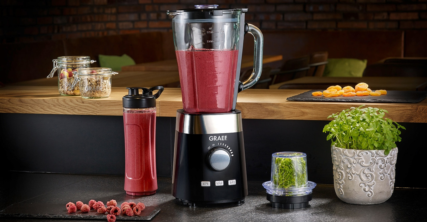Mixer mit Smoothie, Zutaten, Pflanze und Dekoration auf dem Tisch.