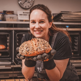Bäckerin hält Brot in Ofenhandschuhen, lächelt in die Kamera, Nahaufnahme, Küche