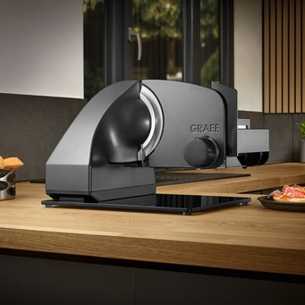 Allesschneider auf Arbeitsfläche aus Holz, Lebensmittel im Hintergrund