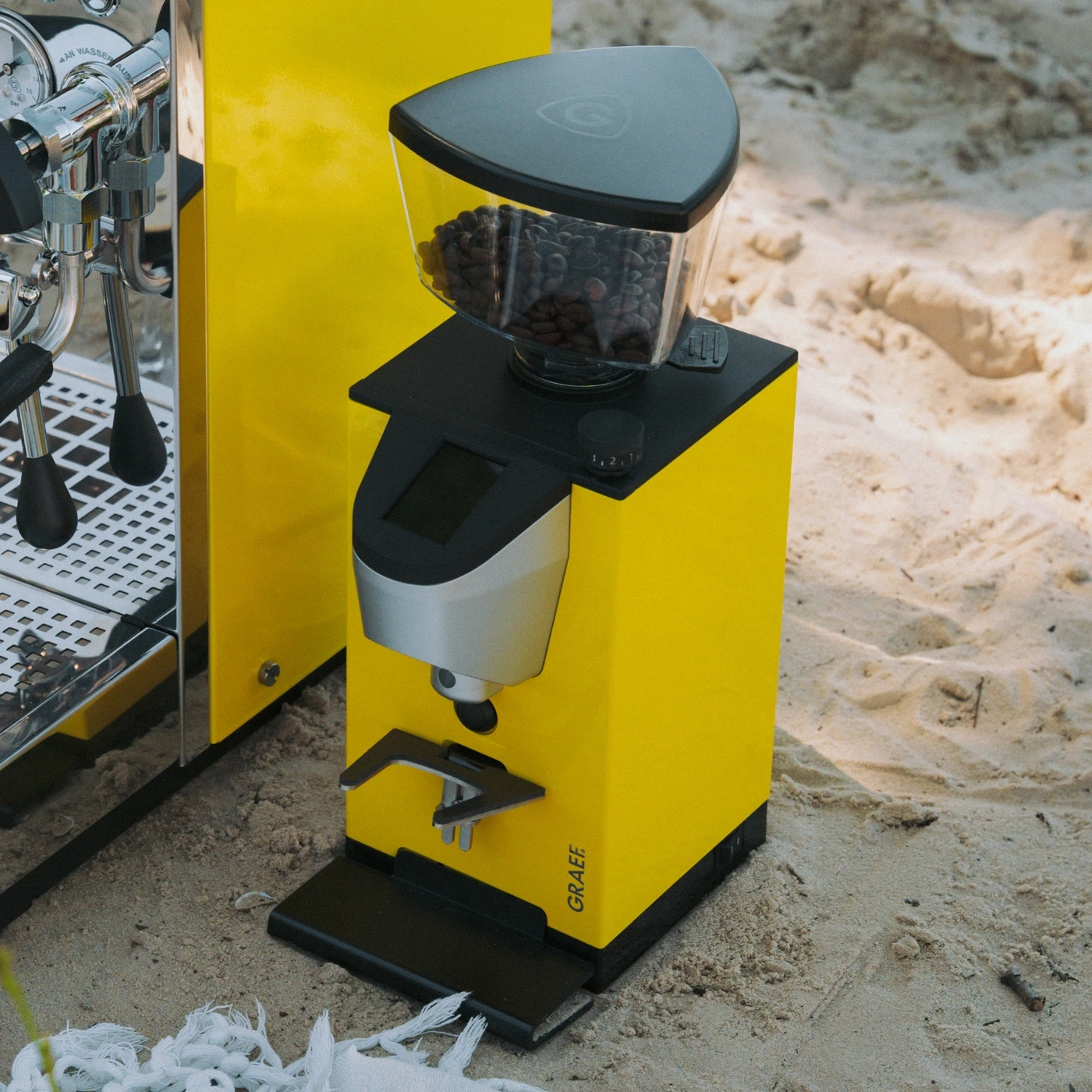 Eine gelbe Kaffeemühle und eine Espressomaschine stehen am Strand im Sand.