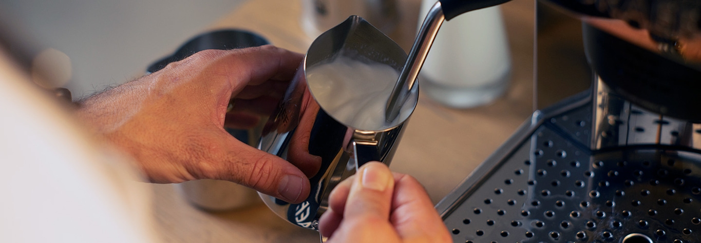 Nahaufnahme einer Person, die Milch in einem Edelstahlkrug aufschäumt, um Kaffee zuzubereiten.