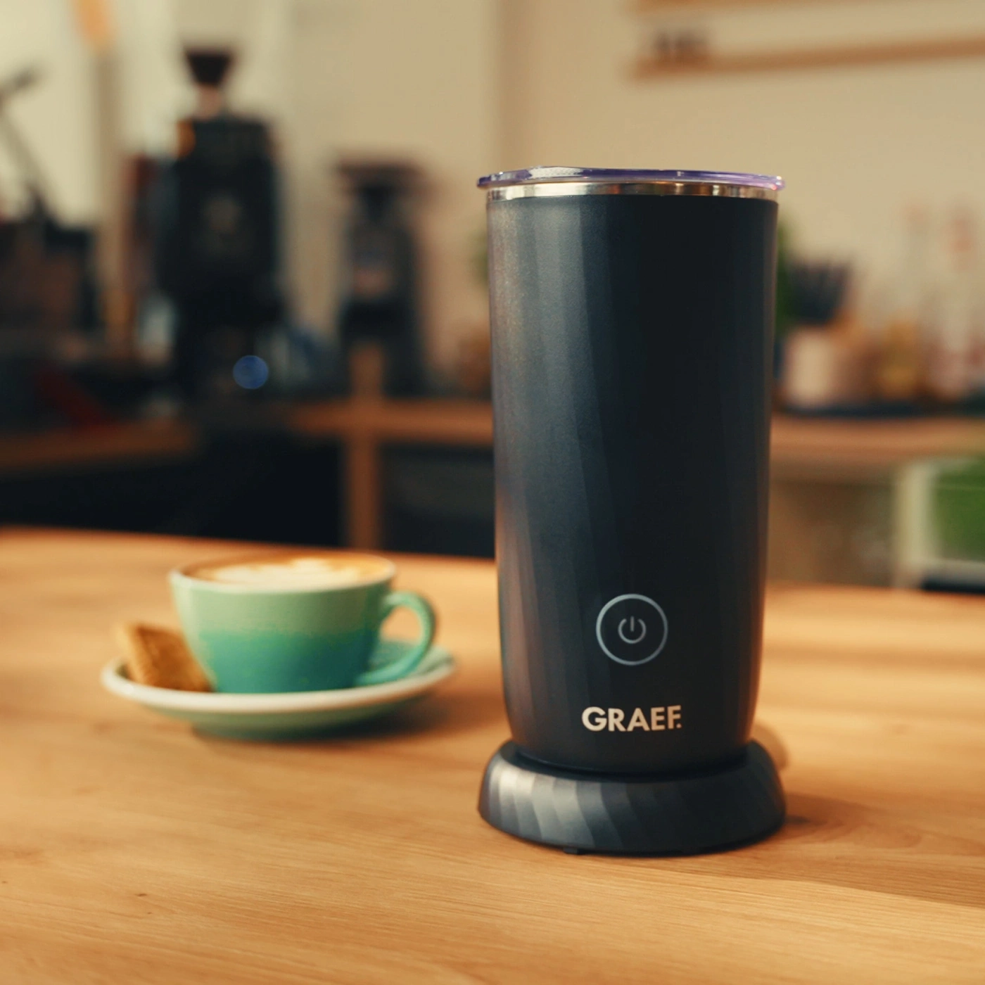 Ein Milchaufschäumer von GRAEF neben einer Tasse Kaffee auf einem Holztisch.