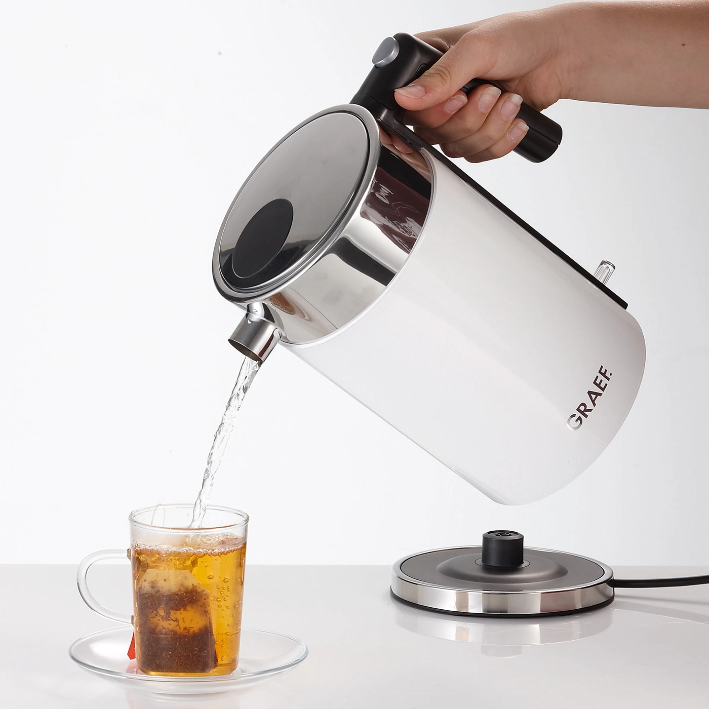 Weißer Wasserkocher gießt heißes Wasser in eine Teetasse mit Teebeutel.