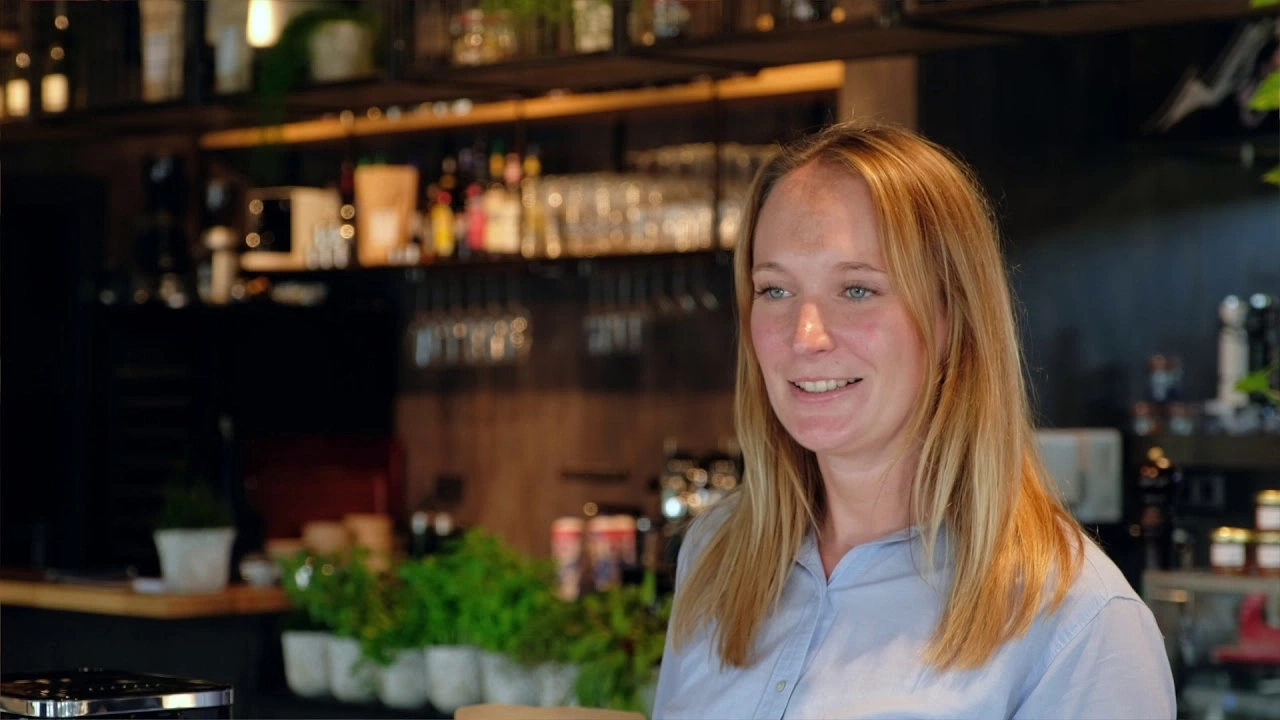 Frau im Café, lächelnd, blondes Haar, blaues Hemd, Hintergrund Bar