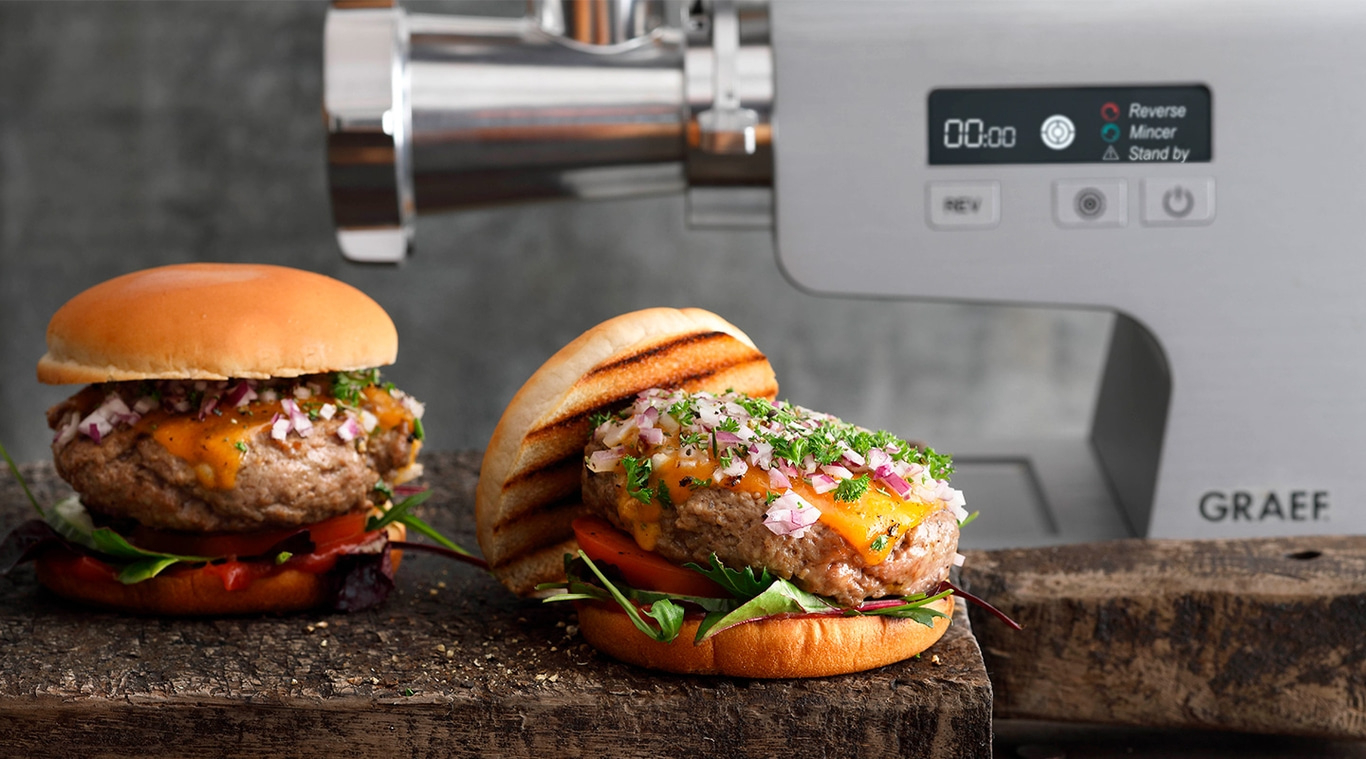 Burger with cheese and onions next to meat grinder, on wooden board