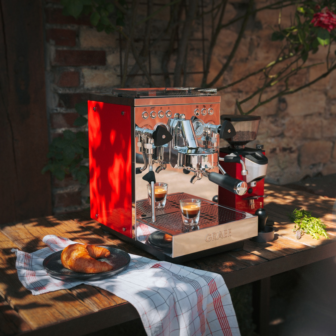 Eine rote Espressomaschine mit zwei Espressoshots, einem Croissant und einer Kaffeemühle im Freien.