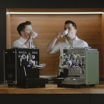 Zwei Männer trinken Kaffee neben Espressomaschinen.