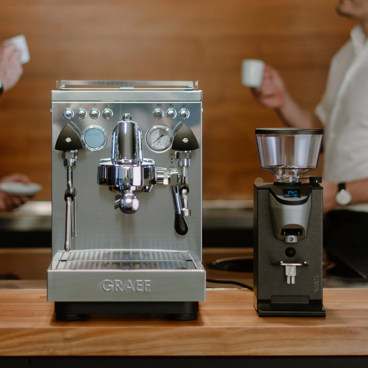 Espressomaschine und Kaffeemühle auf Holzoberfläche, mit Personen im Hintergrund.