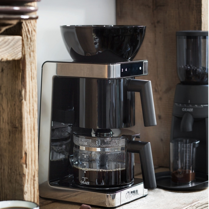 Coffee machine with grinder, glass carafe, black design, wooden surface, Graef brand.