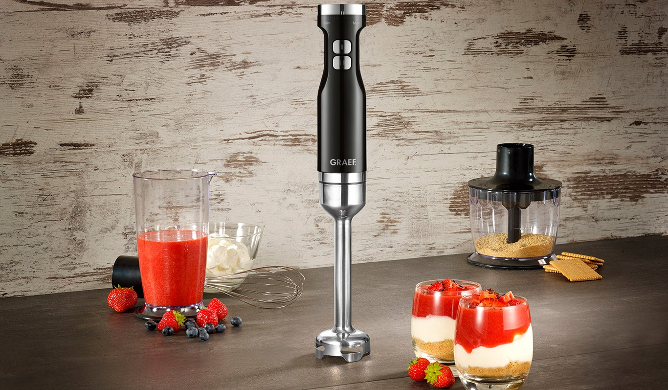 Schwarzer Stabmixer mit Erdbeeren und Dessertgläsern auf dem Tisch.