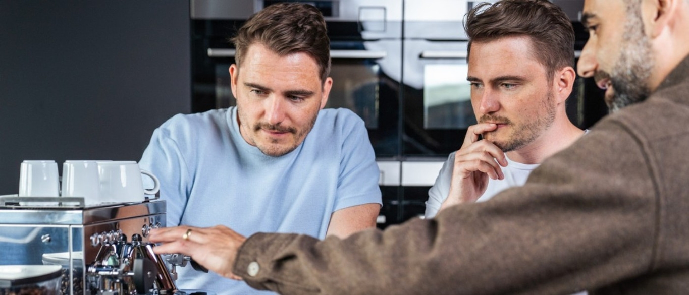 Drei Männer an einer Kaffeemaschine, die sich unterhalten und Kaffee zubereiten.