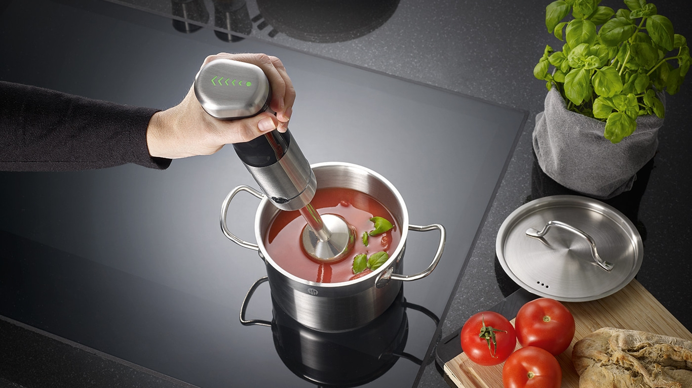 Person püriert Suppe mit einem Stabmixer in einem Topf mit Zutaten.