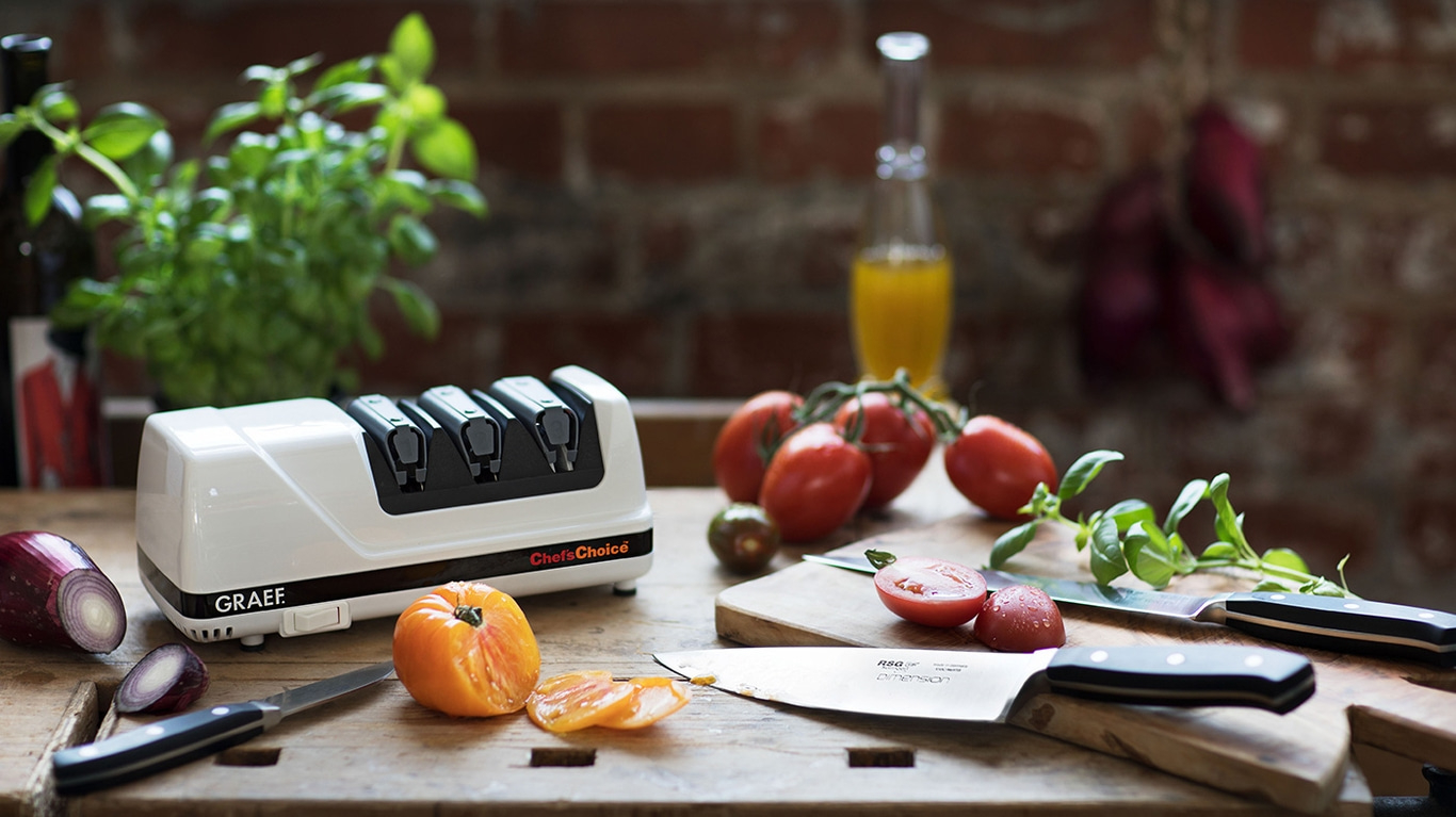 Küchenszene mit Tomaten, Messer, Schleifgerät und Gemüse auf einem Holztisch.