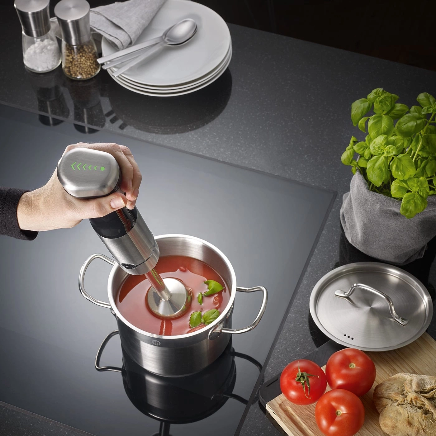 Handmixer in einem Topf mit Tomatensuppe, daneben Tomaten und Basilikum.