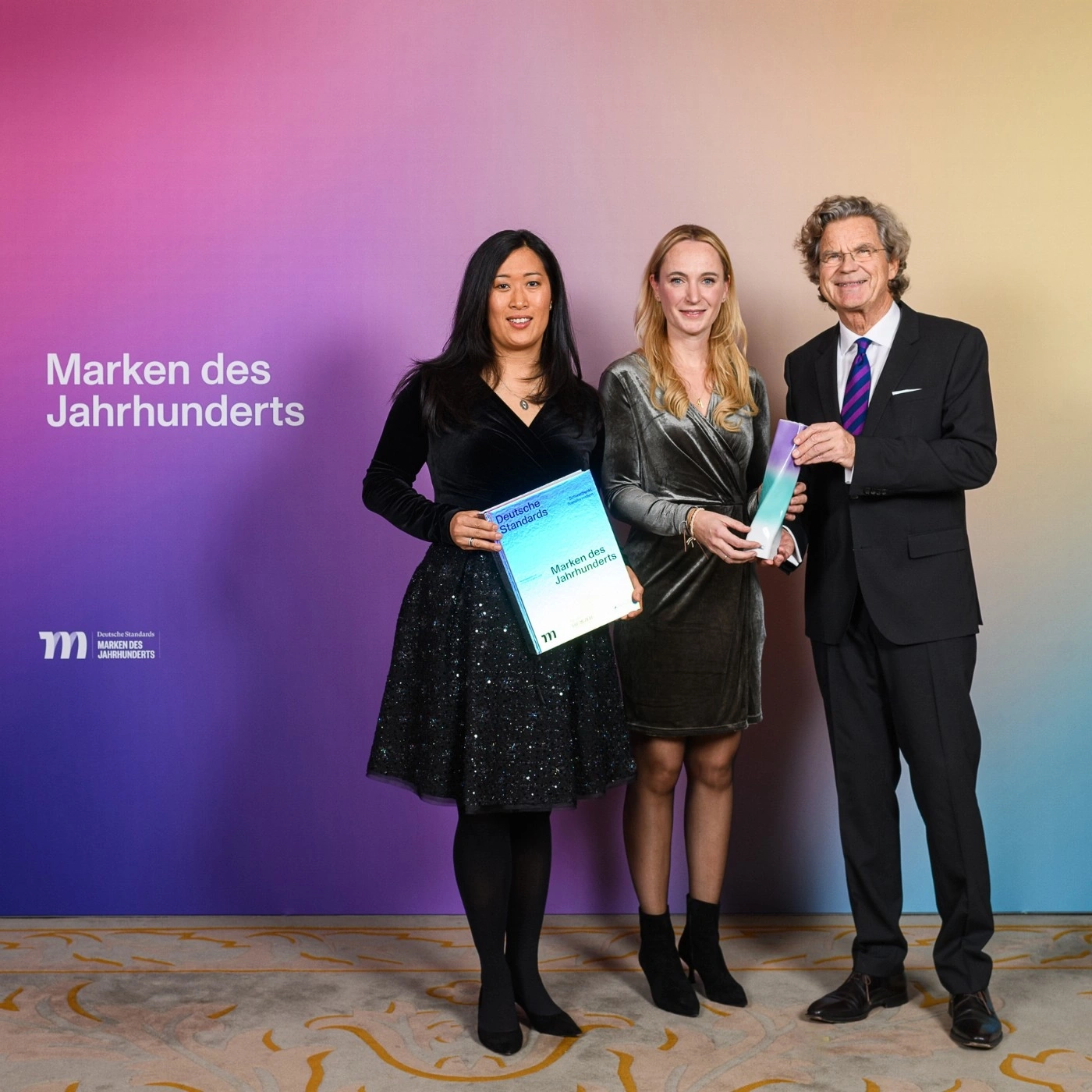 Drei Personen halten Auszeichnungen vor farbigem Hintergrund.