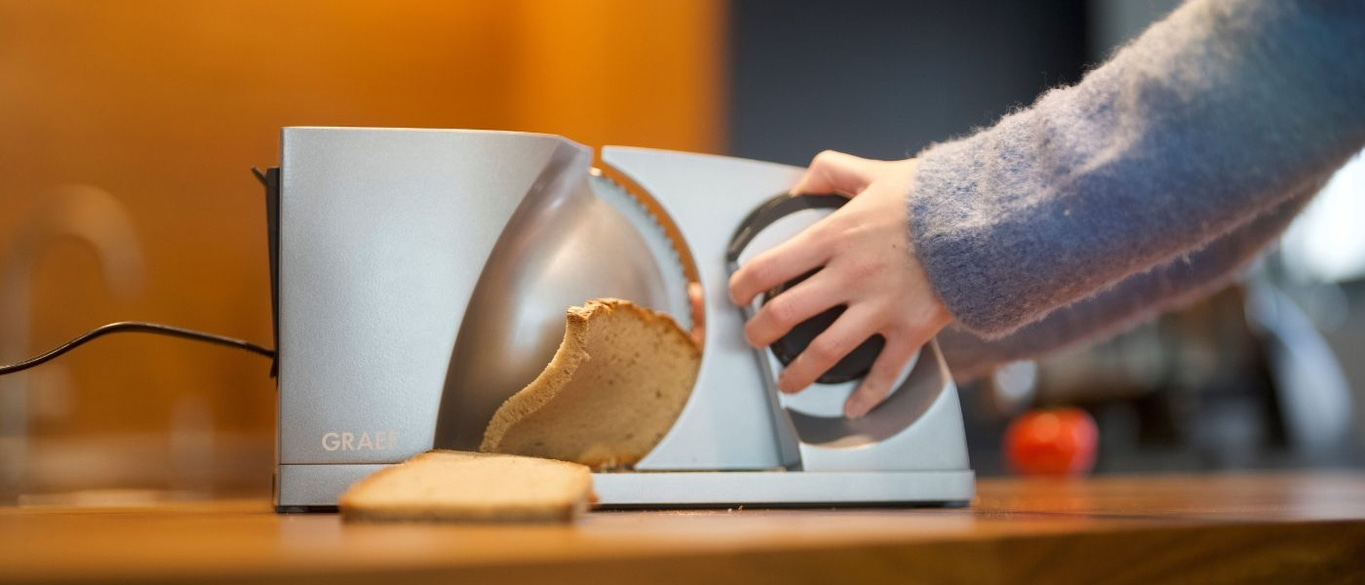 Person cutting bread with a slicer on a wooden surface, close-up shot.