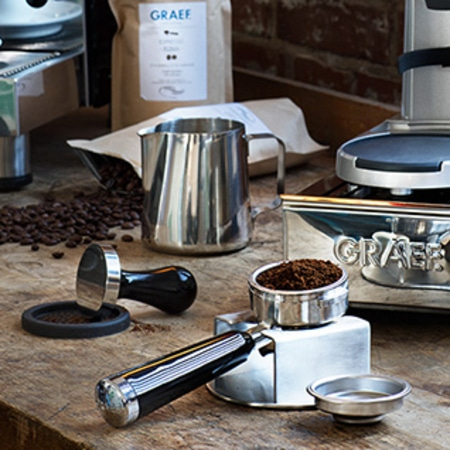 Kaffeemaschine mit Zubehör, Kaffeebohnen und Milchkanne auf Holzoberfläche.