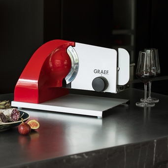 Rote Aufschnittmaschine mit Wurst, Feigen und Weingläsern auf einer Theke.