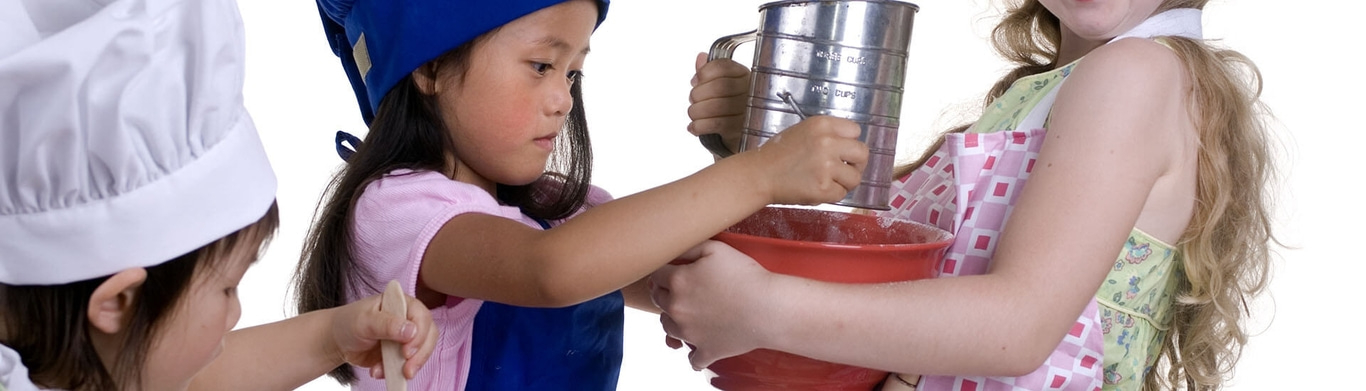 Bambini che cucinano insieme, una ragazza setaccia la farina in una ciotola rossa.
