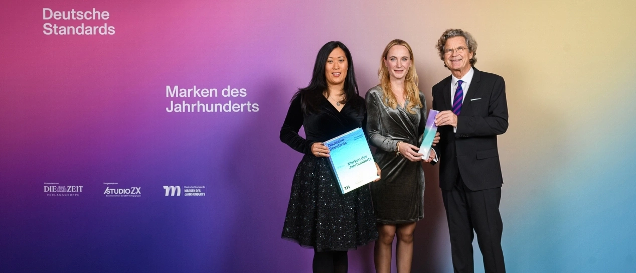 Drei Personen halten Auszeichnungen vor einem farbigen Hintergrund.