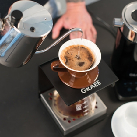 Caffè in preparazione in una tazza, con acqua versata da un bollitore, su un supporto.